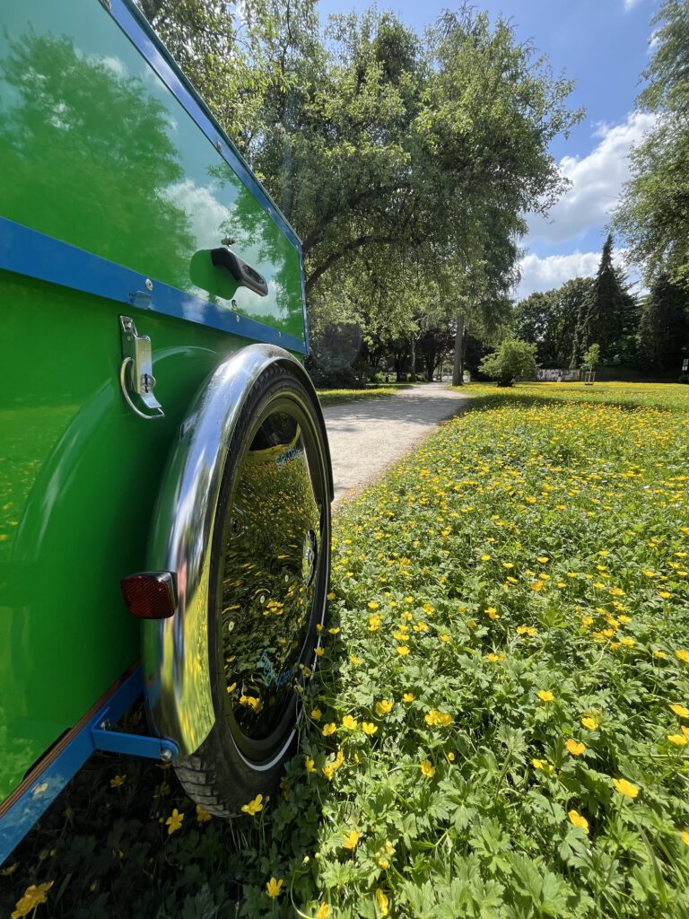 Grünes Lastenfahrrad auf einem Weg im Stadtpark in Moers, in einem Meer von Butterblumen, umgeben von Bäumen, die Sonne scheint und der Himmel ist blau mit wenigen Wolken.